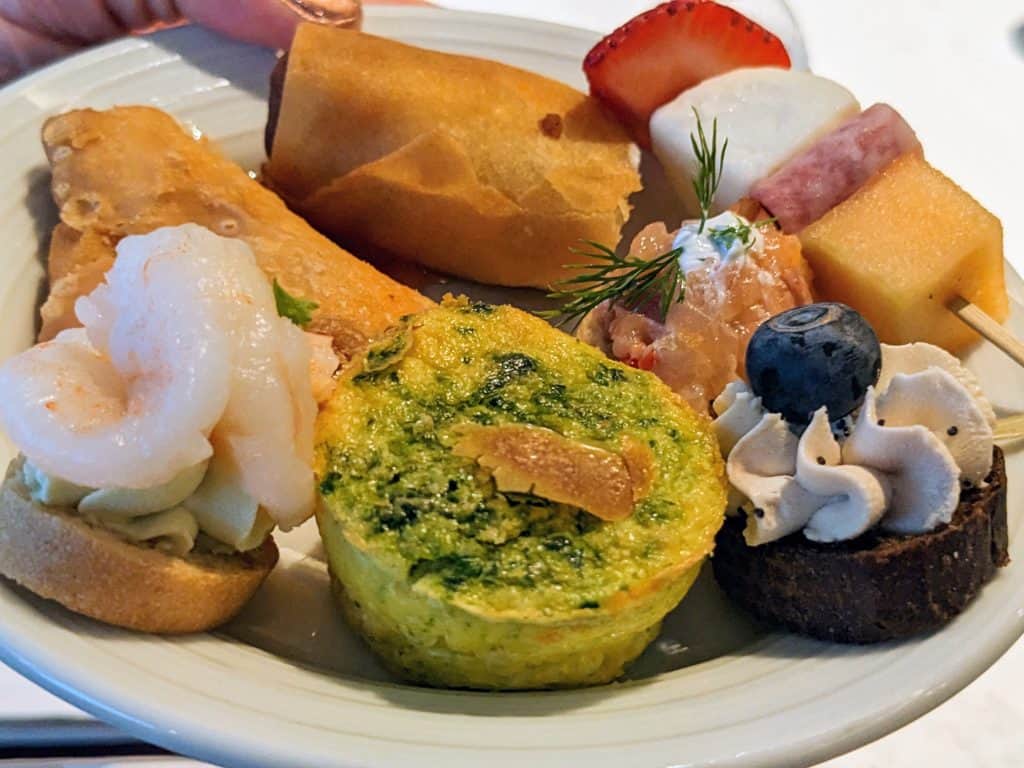 Plate of finger foods including mini spinach quiche; mini egg roll; mini samosa; kebab with strawberry, mozzarella, salami, and cantaloupe, salmon and dill canape; blueberry and cream cheese canape; shrimp and cream cheese canape