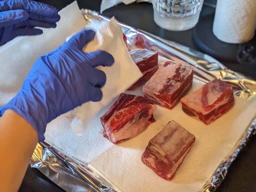 Raw beef short ribs lying on a paper towel lined pan while being patted dry with more paper towels