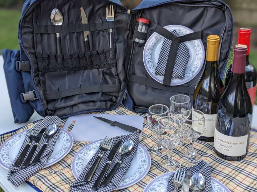 Picnic backpack with picnic plates, napkins, utensils, glasses, a small cutting board, bottles of wine, and a wine opener