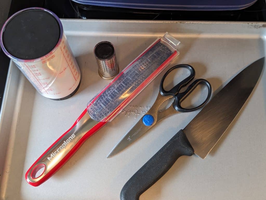 Kitchen tools including a chef's knife, kitchen shears, a microplane zester, a large adjustable measuring cup, and a small adjustable measuring cup