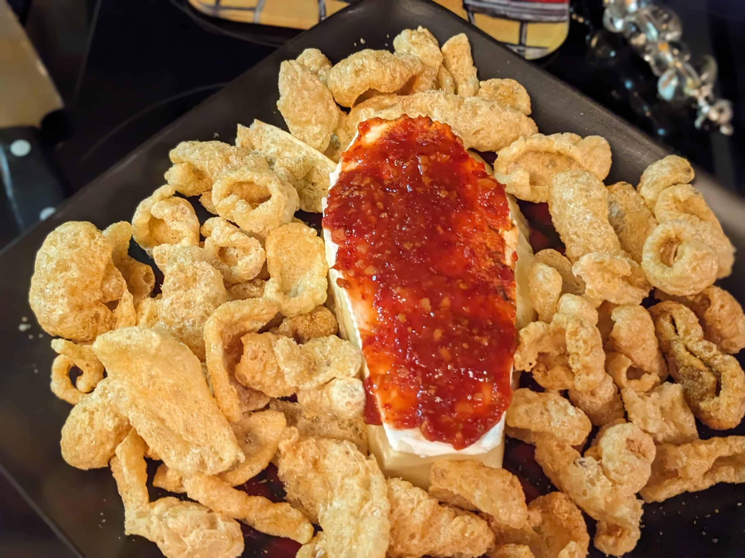 Brie cut in a coffin shape and covered with chili garlic sauce on a plate surrounded by pork rinds