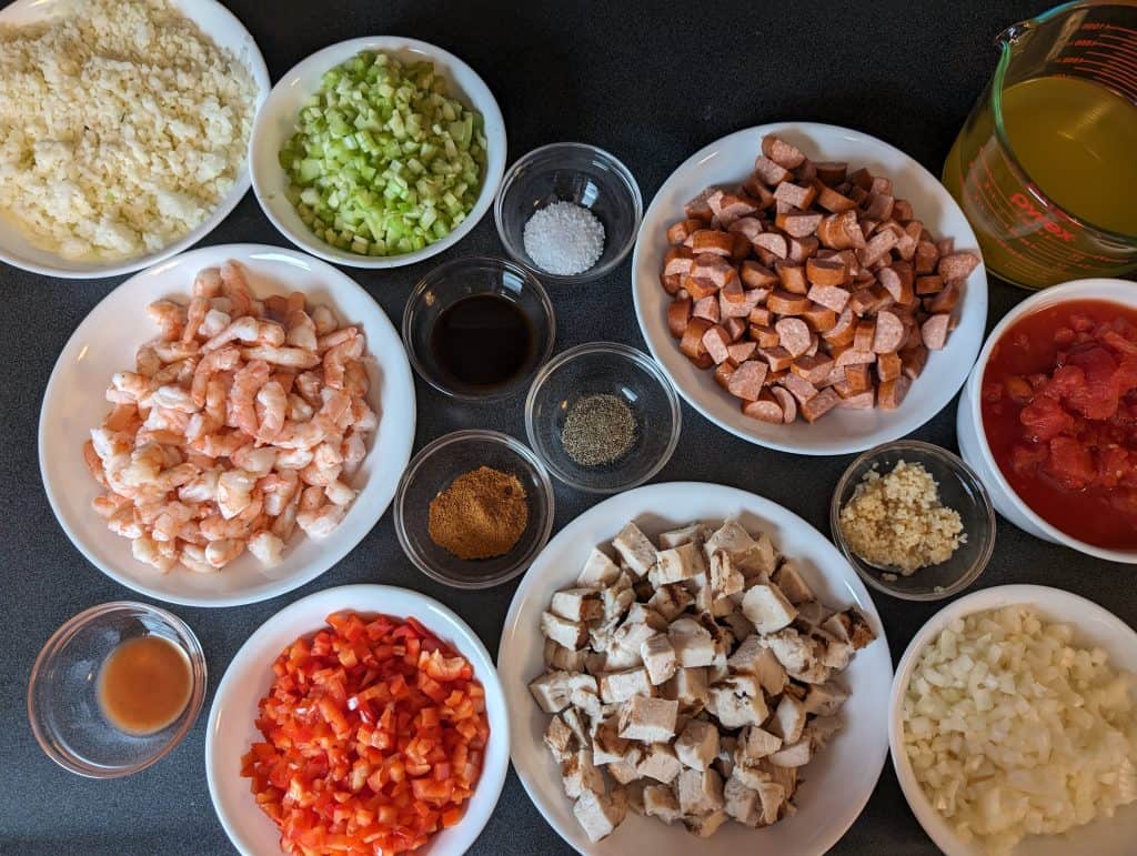 Ingredients for Low Carb Jambalaya - (clockwise, circling inward) cauliflower rice, diced celery, salt, andouille sausage, chicken broth, diced tomatoes, diced onions, cubed chicken pieces, diced bell pepper, hot sauce, chopped shrimp, Worcestershire sauce, black pepper, minced garlic, Creole seasoning