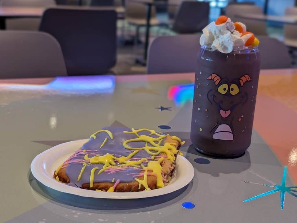 Blueberry filled pastry tart with purple and yellow icing on a plate next to a grape smoothie in a plastic glass with a dragon face on it topped with whipped cream and freeze-dried skittles