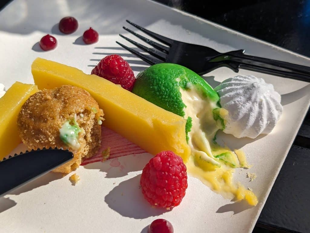 A Deconstructed Key Lime Pie consisting of meringues, a lime custard shaped and colored like a lime, fresh raspberries, gelatinized lime curd, a mini cake, and dollops of raspberry coulis partially eaten