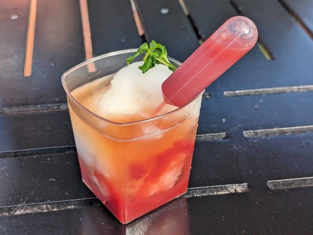 A Deconstructed Strawberry Mint Julep in a plastic rocks glass made with mint sorbet, strawberry puree, and bourbon with part of the strawberry puree in a little decorative syringe stuck into the mint sorbet