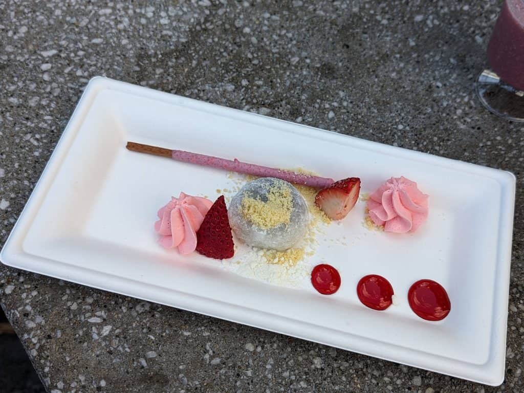 A plate with a strawberry filled mochi, 2 strawberry pieces, dollops of strawberry whipped cream and strawberry glaze, and a strawberry Pocky stick