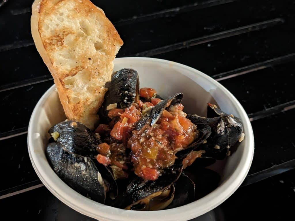 A bowl filled with-Roasted Mussels, Sautéed Tomatoes, Garlic and Fresh Herbs with a slice of toasted bread