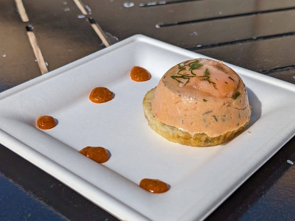 A plate containing a smoked salmon mousse on an herbed blini topped with an herbed aspic next to dollops of a red pepper coulis