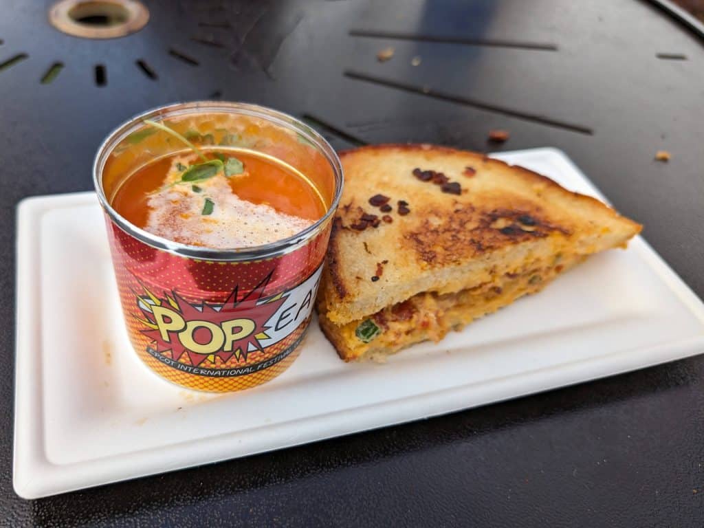 A decorative can of tomato soup plated next to a grilled sandwich with pimento cheese and fried green tomatoes