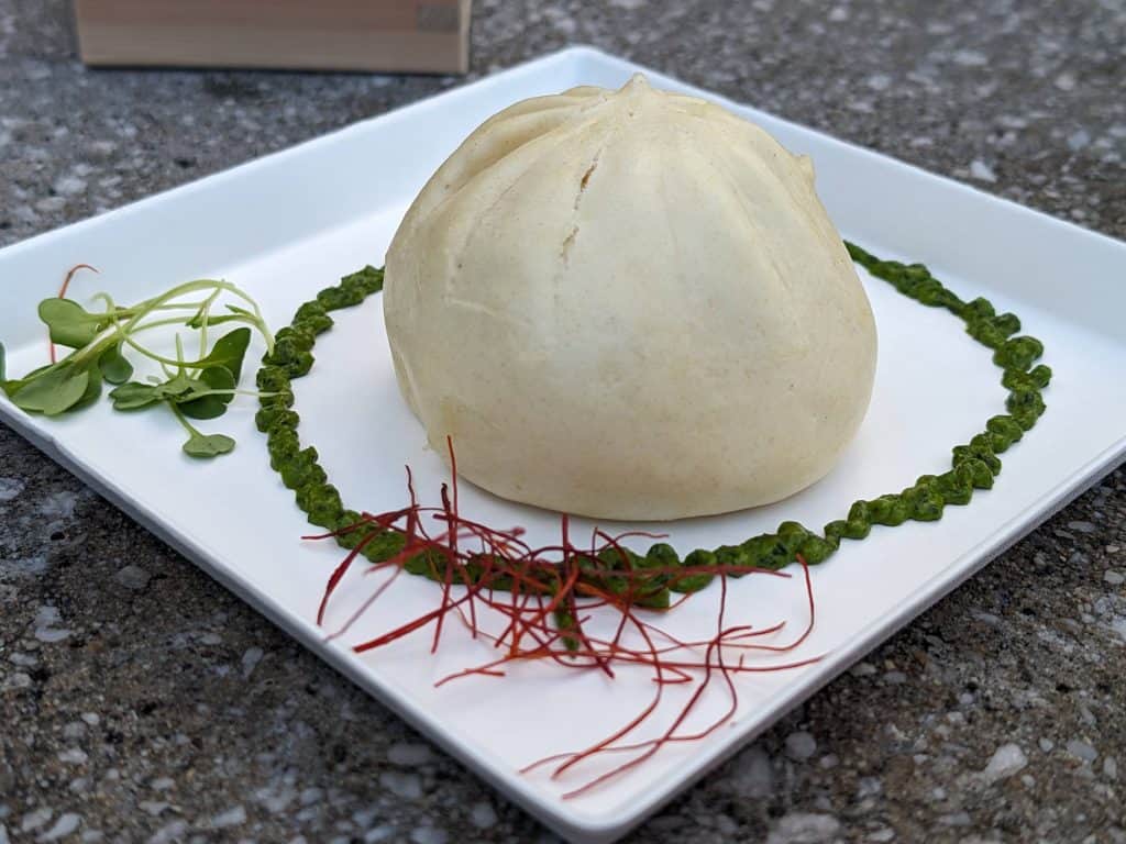 A large steamed bun filled with ground Waygu beef on a plate circled by green shiso sauce