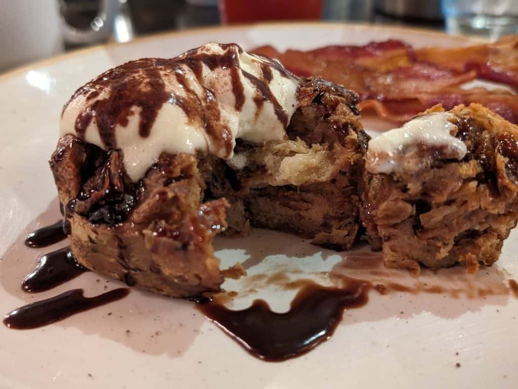 A cross-section view of Tiramisu Bread Pudding topped with a sweet cheese sauce and drizzled with chocolate with bacon in the background