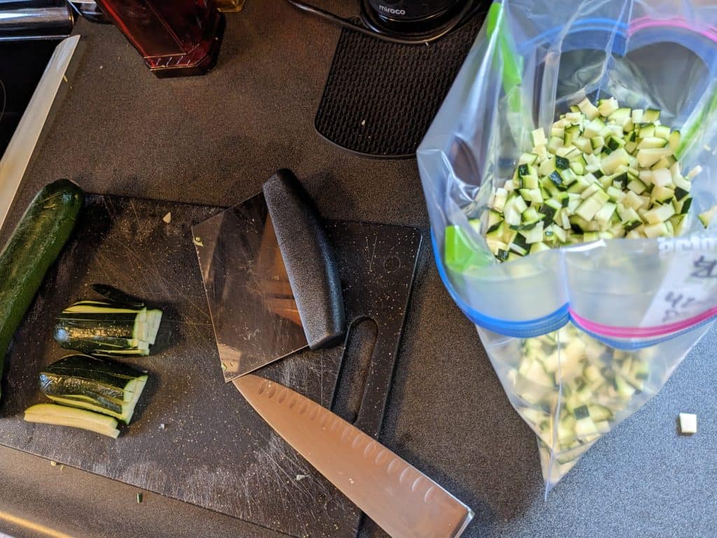 Zucchini being diced on cutting board and a gallon zip top bag being held open to store the diced zucchini