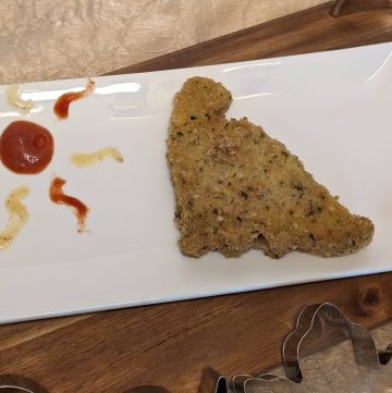 Gluten-Free Dinosaur Chicken Patty on a plate with dabs of Sugar Free Honey Mustard Dipping Sauce and No Sugar Added Ketchup on the plate next to it in the shape of a sun