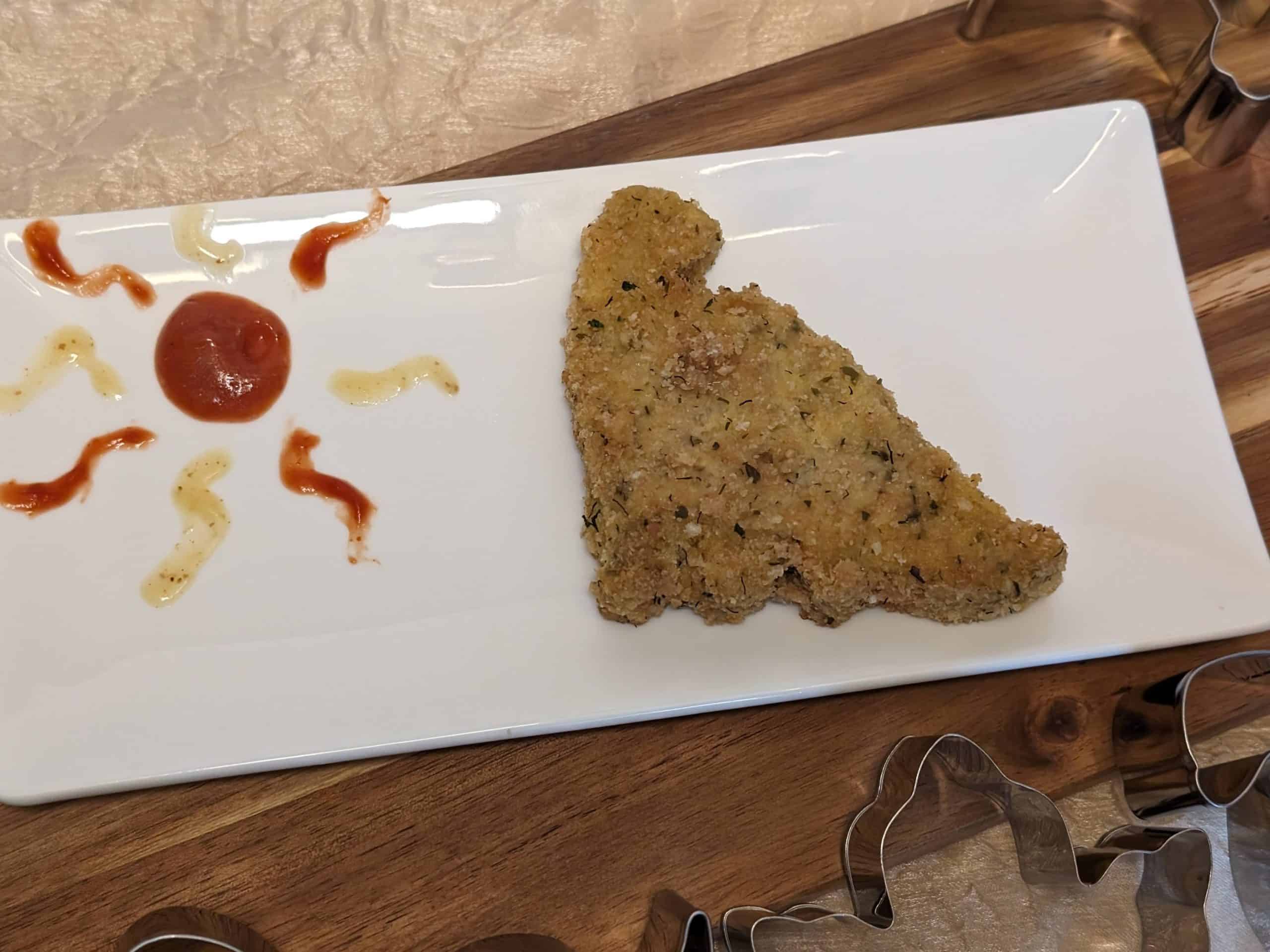Gluten-Free Dinosaur Chicken Patty on a plate with dabs of Sugar Free Honey Mustard Dipping Sauce and No Sugar Added Ketchup on the plate next to it in the shape of a sun