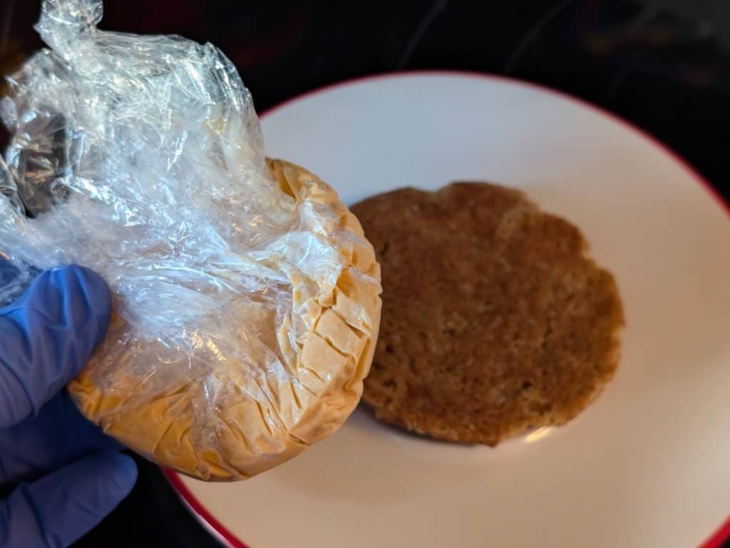 A frozen disk of Chipotle Keto Ice Cream in plastic wrap next to a plate with an upside down Keto Snickerdoodle Cookie as the 2nd step in constructing a Keto Chipotle Ice Cream Sandwich