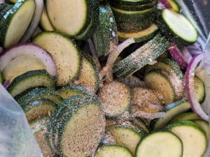 Sliced zucchini and red onion in a zip top bag with seasoning sprinkled on top of them