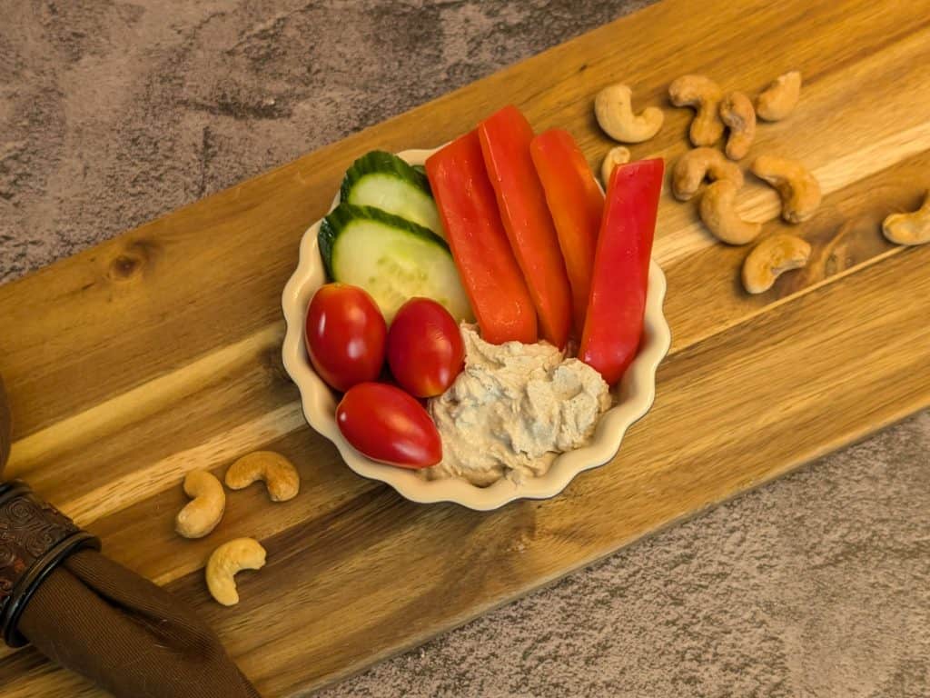 Creamy Za'atar Cashew Dip in a dish with slices of cucumber, red bell pepper, and grape tomatoes with some cashews on the side