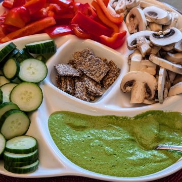 Garlicky Spinach Cashew Dip in a serving dish with sliced cucumbers, mushrooms, and red bell peppers as well as flaxseed crackers