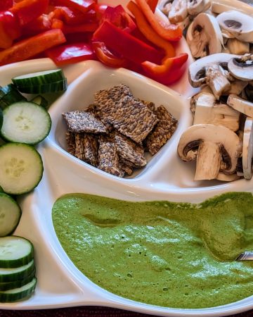 Garlicky Spinach Cashew Dip in a serving dish with sliced cucumbers, mushrooms, and red bell peppers as well as flaxseed crackers