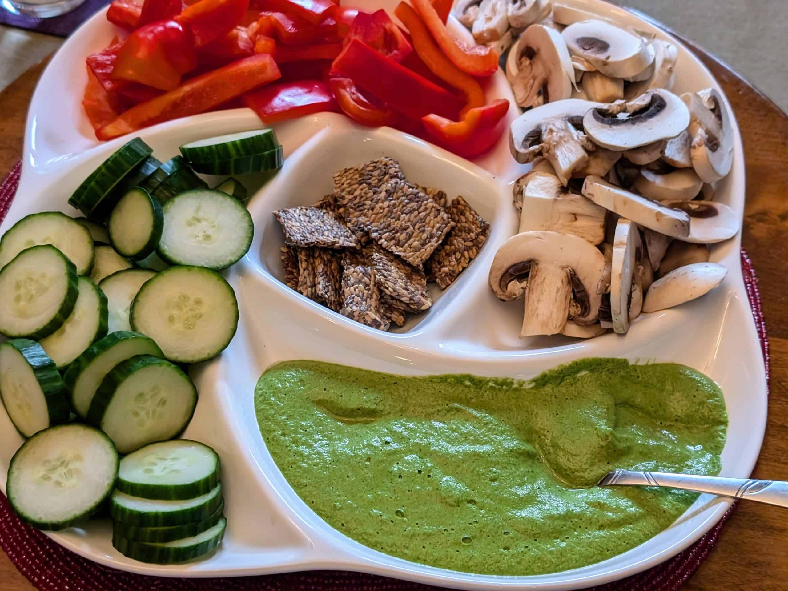 Garlicky Spinach Cashew Dip in a serving dish with sliced cucumbers, mushrooms, and red bell peppers as well as flaxseed crackers