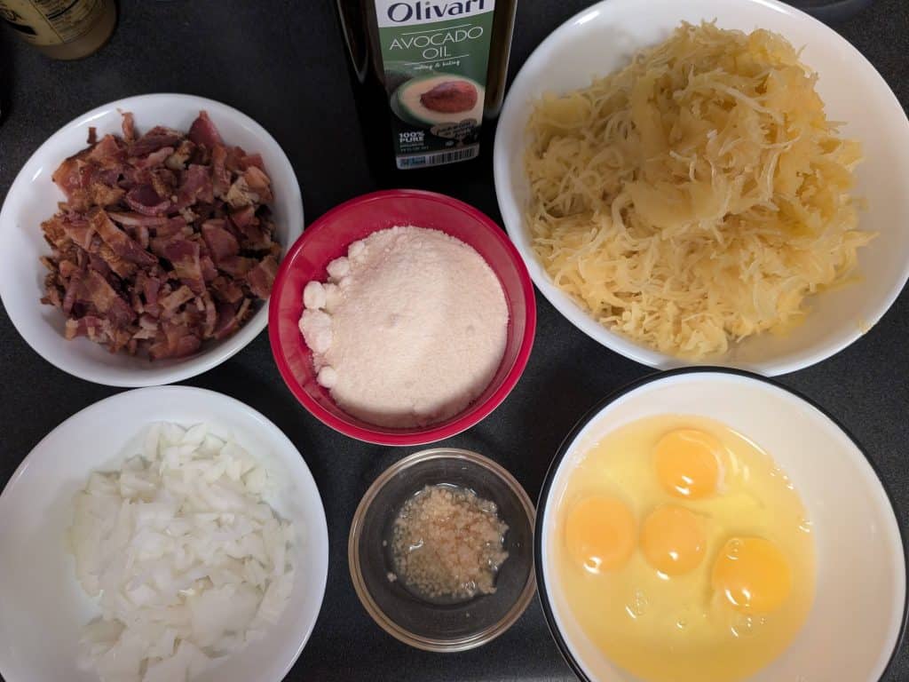 Ingredients for Spaghetti Squash Carbonara, including cooked bacon pieces, grated parmesan cheese, chopped onion, minced garlic, eggs, avocado oil, and shredded spaghetti squash