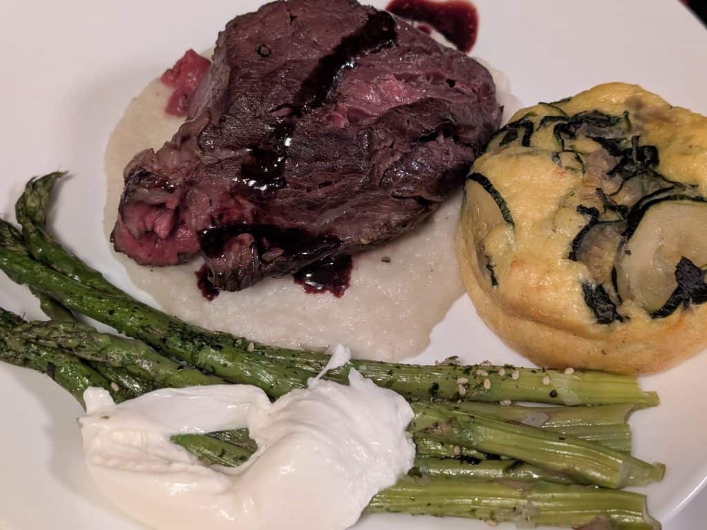 A white plate containing a slice of Beef Tenderloin on a bed of Turnip and Rosemary Purée with a drizzle of Blackberry Red Wine Reduction Sauce, next to an individual-size Zucchini and Gouda Soufflé and Roasted Asparagus topped with Furikake and Burrata