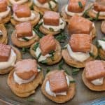A serving plate filled with Savory Blini Salmon Bites. Each savory blini is topped with a dollop of creme fraiche and a cube of smoked salmon with fresh chives sprinkled on top.