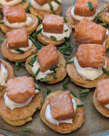 A serving plate filled with Savory Blini Salmon Bites. Each savory blini is topped with a dollop of creme fraiche and a cube of smoked salmon with fresh chives sprinkled on top.