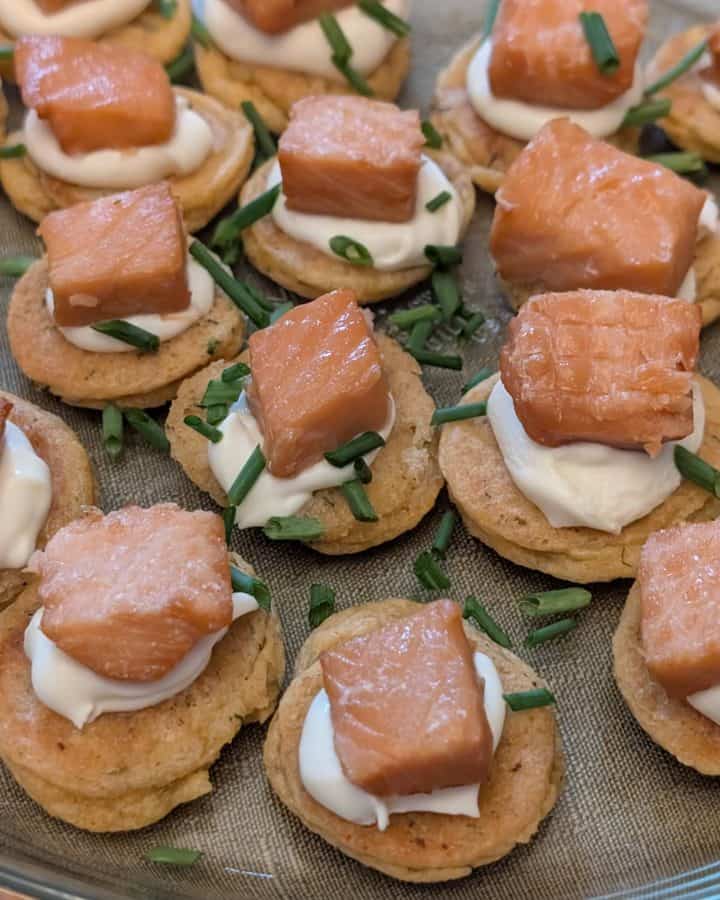 A serving plate filled with Savory Blini Salmon Bites. Each savory blini is topped with a dollop of creme fraiche and a cube of smoked salmon with fresh chives sprinkled on top.