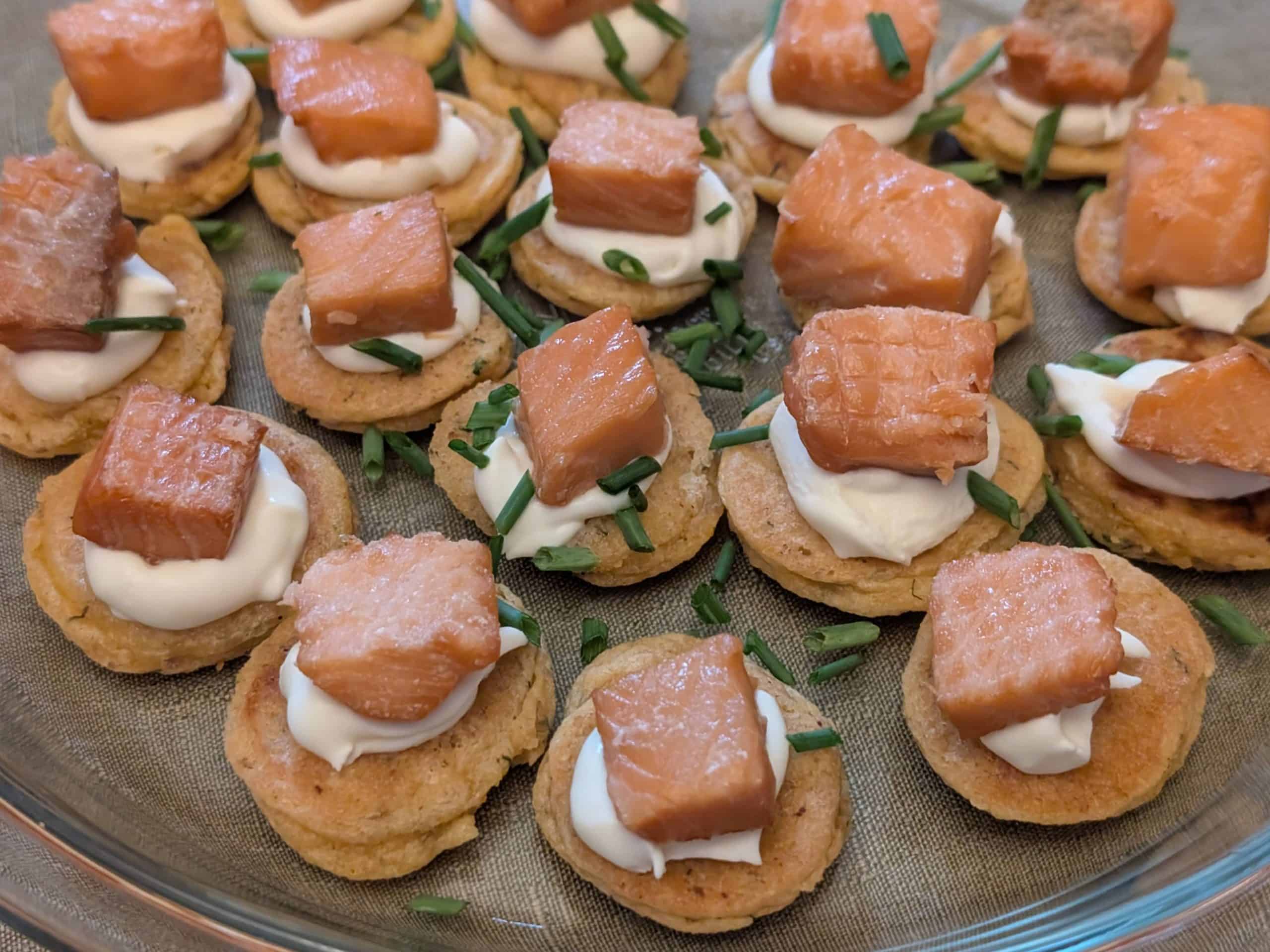 A serving plate filled with Savory Blini Salmon Bites. Each savory blini is topped with a dollop of creme fraiche and a cube of smoked salmon with fresh chives sprinkled on top.