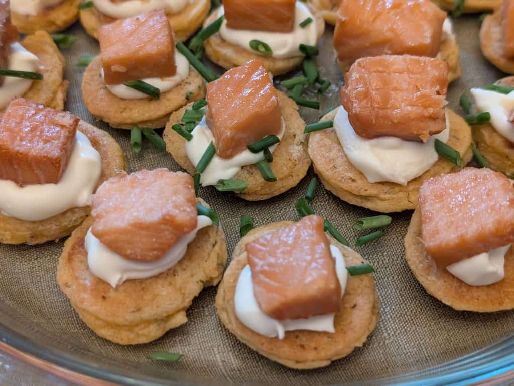 A serving plate filled with Savory Blini Salmon Bites. Each savory blini is topped with a dollop of creme fraiche and a cube of smoked salmon with fresh chives sprinkled on top.