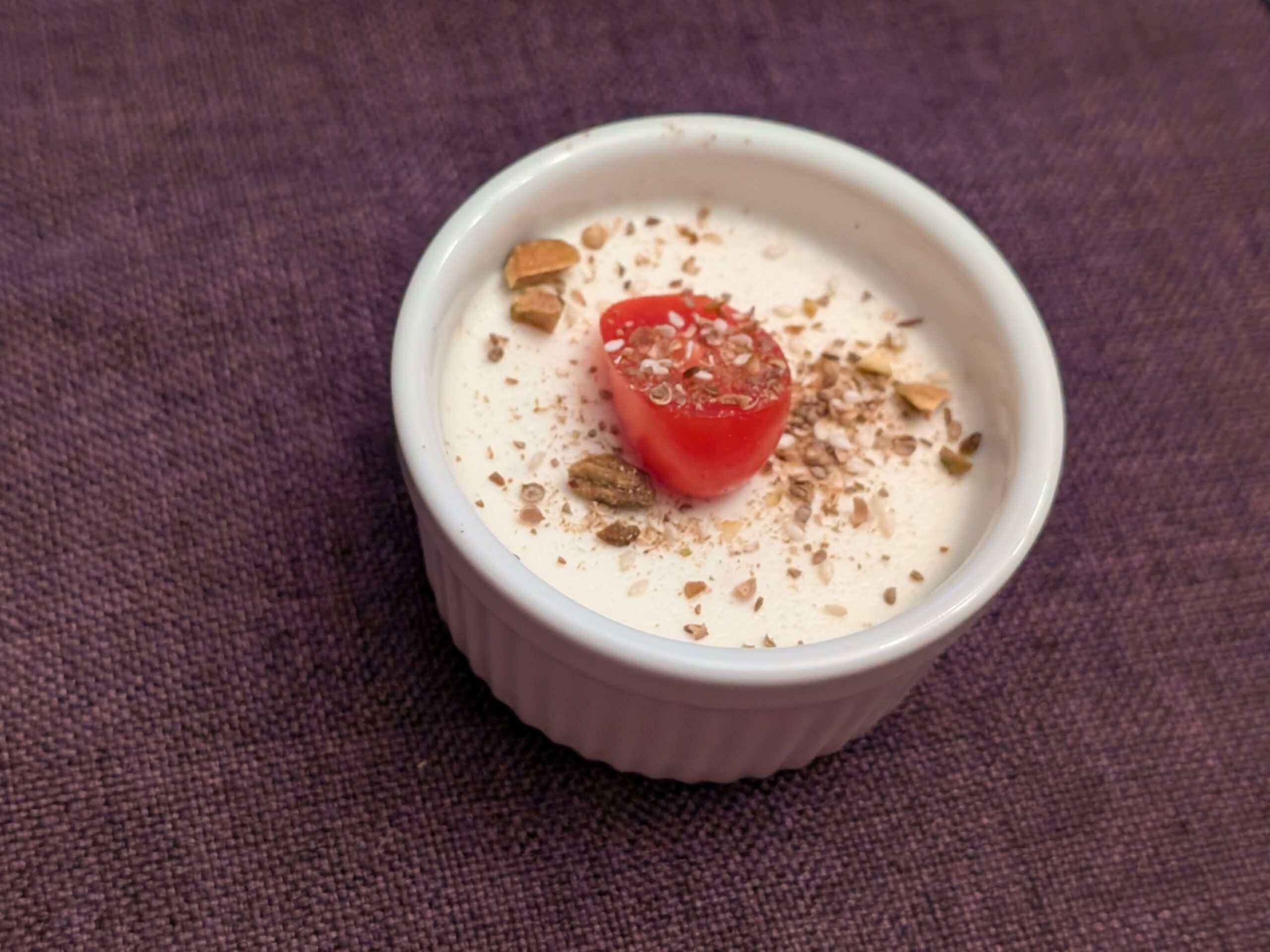 Savory Goat Cheese Panna Cotta in a small, white ramekin topped with Dukkah and a sliced grape tomato