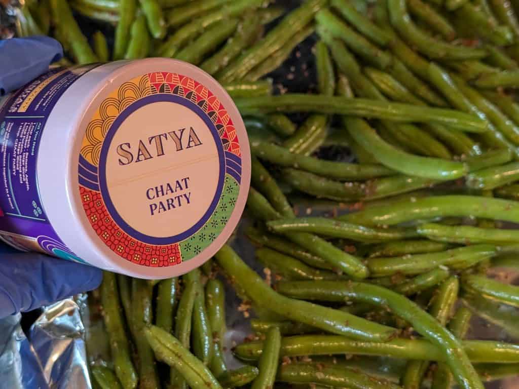 Raw green beans seasoned with Chaat Party Spice Blend on a sheet pan ready to roast with spice tin in picture