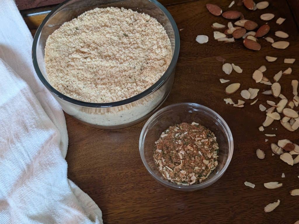 A bowl of Dinner Party Diaries Savory Baking Mix and a spice blend in a prep bowl on a wooden tray sprinkled with almonds